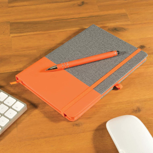 A grey notebook with an orange cover and an orange pen placed on a wooden desk beside a keyboard and mouse.