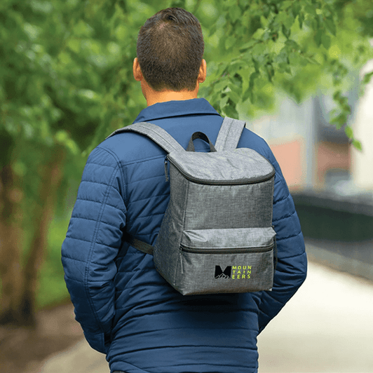 A grey backpack cooler with a logo, worn on the back of a person outdoors, surrounded by greenery.