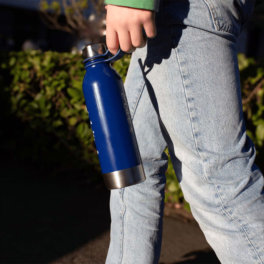 A drink bottle in blue stainless steel, featuring a handle and metallic base, held by a person in denim.