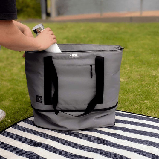 A grey cooler bag is being filled with a drink bottle, set on a striped blanket on grass.