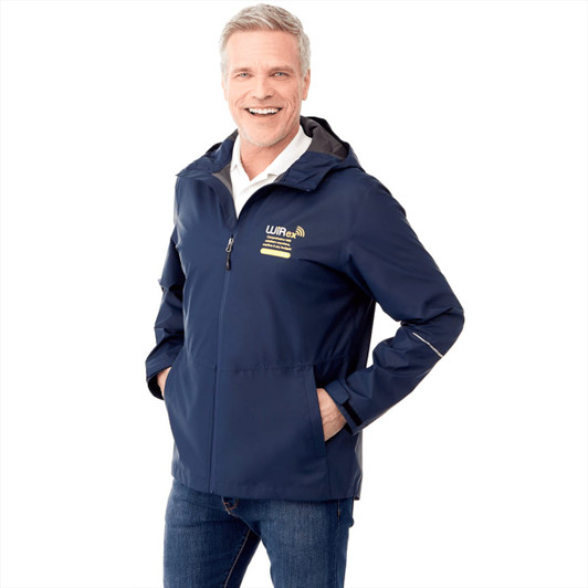 A man in a navy rain jacket with a hood and a logo, smiling and standing with hands in pockets.