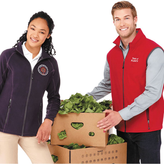 A man in a red vest stands beside a woman in a dark purple vest, both holding boxes filled with leafy greens.