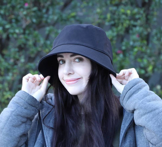 A black bucket hat featured on a model with long dark hair, set against a green backdrop. The hat has a logo on it.