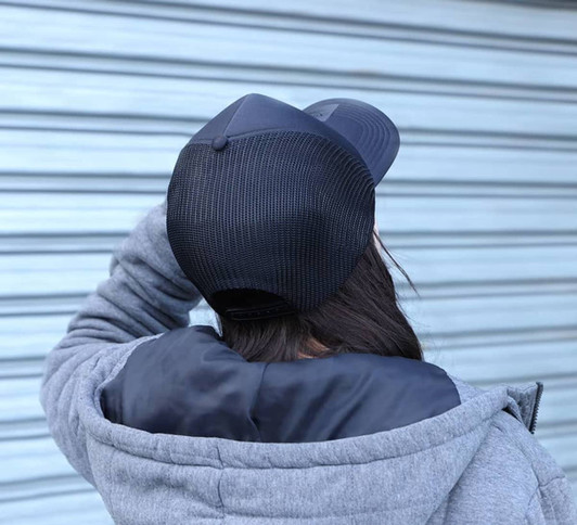 A black trucker cap with a mesh back, worn by a person in a grey hoodie. The background features a blue-grey wall.