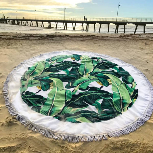 A round towel featuring green banana leaves on a white background, set against a sandy beach with a pier in the background.