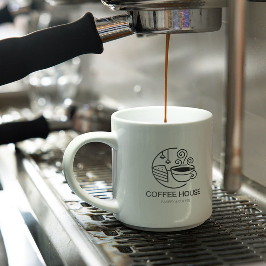 A stackable white coffee mug with a logo, placed under an espresso machine as coffee is being poured.