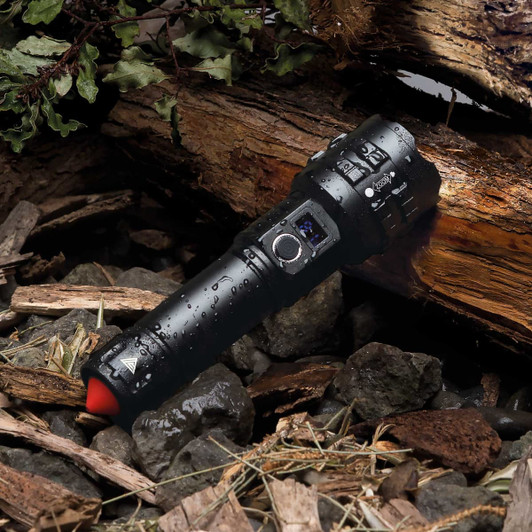 A black waterproof torch lies on a bed of rocks and wood, with droplets of water on its surface.