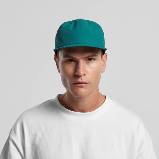 A teal cap with a flat brim and a fitted design, worn by a model in a white sweatshirt.