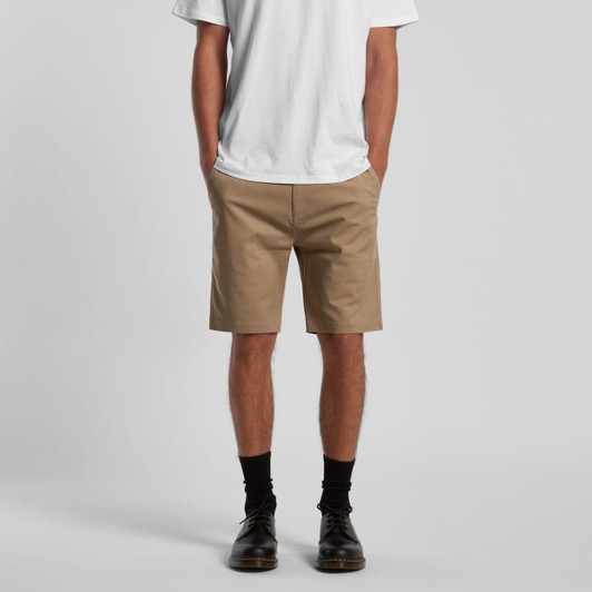 Tan dress shorts displayed on a model wearing a white t-shirt and black shoes. The shorts feature pockets and a logo.