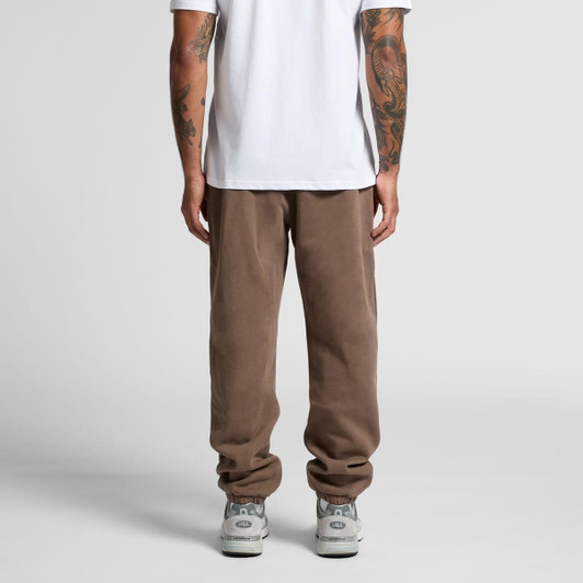 Brown track pants with a relaxed fit, featuring tapered legs and elastic cuffs. The model wears a plain white t-shirt.