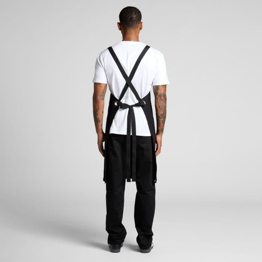 A black apron with crossed straps, featuring a logo, worn over a white T-shirt and black pants.