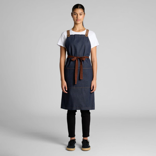 A denim apron featuring brown leather straps and a tie, displayed on a model against a neutral background.