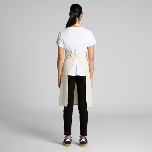 A linen apron in a natural beige colour, worn over a white T-shirt and black pants, showing the back view.