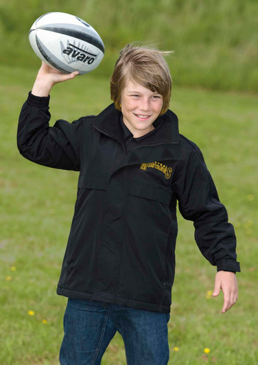 A youth wearing a black rain jacket, holding a rugby ball, standing on grass outdoors. The jacket has a logo.