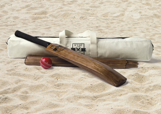 A cricket bat, two wooden stumps, a red cricket ball, and a cream carry bag with a logo, set on sand.