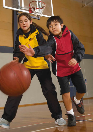 A boy in a red rain jacket and a girl in a yellow rain jacket play basketball indoors. Both jackets have branding.