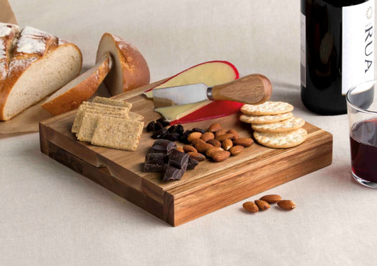 A wooden clamshell cheese board features cheese, crackers, nuts, chocolate, and a knife, alongside bread and wine.