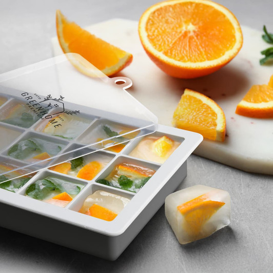 A Blizzard Ice Tray with Lid in white, featuring citrus and herb ice cubes; a sliced orange is nearby.