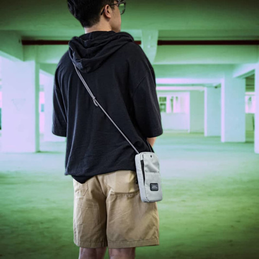 A grey crossbody bag with a front pocket, displayed on a person wearing a black hoodie and shorts.