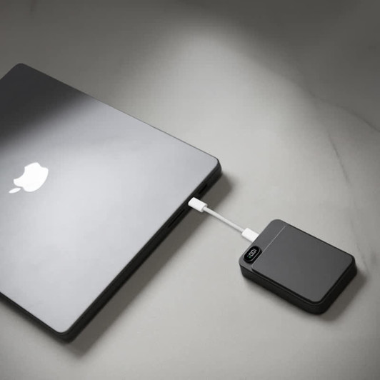 A black magnetic wireless power bank connected to a laptop on a light surface. The power bank has a logo.