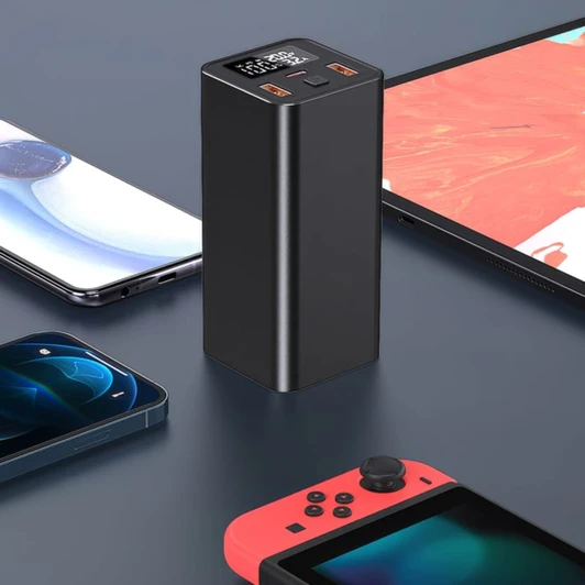A black power bank stands upright, surrounded by smartphones and a gaming console on a dark surface.