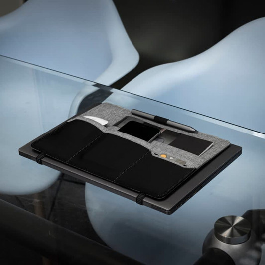 A grey and black clipboard with pockets, a pen, and a phone holder, placed on a glass table.