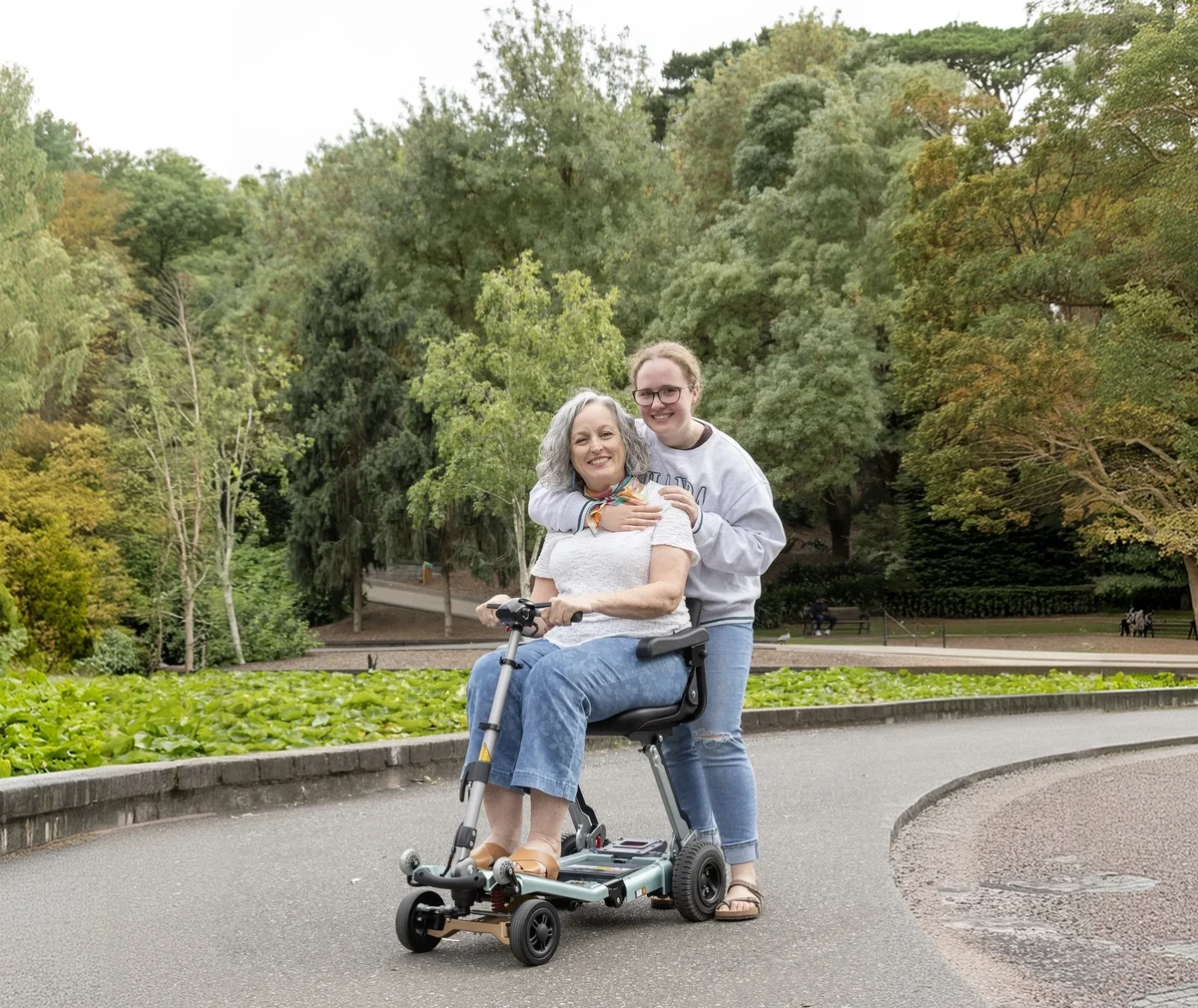 Luggie owner seated on her scooter in a park with a family member standing behind her