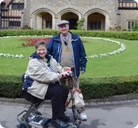 Couple in front of a garden and manor house, one seated on a Luggie