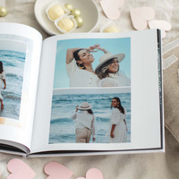 Open hardcover photo book with beachside portraits of two women in white clothing, surrounded by white and gold chocolates and pink paper hearts