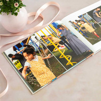 Open hardcover photo book showing a child playing on a colorful playground, styled with a vase of flowers and ribbon.