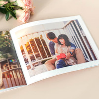 Open softcover photo book featuring a couple sitting on a balcony at sunset, with one partner holding a red gift box, placed beside pale pink roses