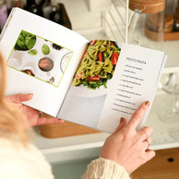 Open softcover photo book featuring a custom recipe layout with pesto pasta image and ingredient list, held in kitchen setting.