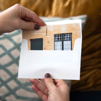Horizontal card with a photo of a yellow brick building being inserted into a white envelope, held in two hands
