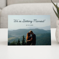 Horizontal wedding announcement card on a white table, showing a couple embracing on a mountaintop with the text “We’re Getting Married!”
