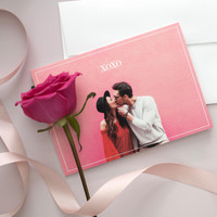 Folded Valentine's Day card featuring a couple kissing in front of a pink wall with the text “XOXO,” styled with a single pink rose, soft ribbon, and matching envelope