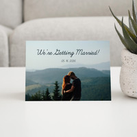 Folded save the date card featuring couple portrait with mountain backdrop and wedding date text, displayed on tabletop beside potted plant.