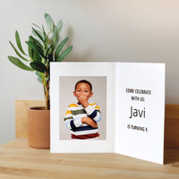 Open folded birthday invitation card with child portrait and “Come celebrate with us” message, displayed beside potted plant on wood table.