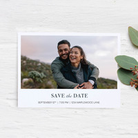 Flat save the date card with landscape couple portrait and event details, printed on white cardstock and styled on a light wood surface with greenery.