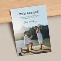 Flat engagement announcement card featuring proposal photo on lakeside dock with headline text and couple names, displayed on neutral tabletop.