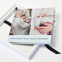 Layflat photo book with newborn collage cover titled “Hello World: Henry James Alexander,” displayed inside a white keepsake box with black ribbon closure.