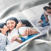 Layflat photo book open to a full-page family portrait of a mother hugging two children indoors, displayed on soft fabric with ribbon accents.