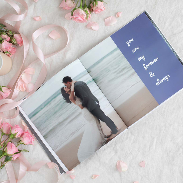 Open hardcover photo book showing a couple embracing on a beach with the text “you are my forever & always” on the opposite page, surrounded by roses and ribbon
