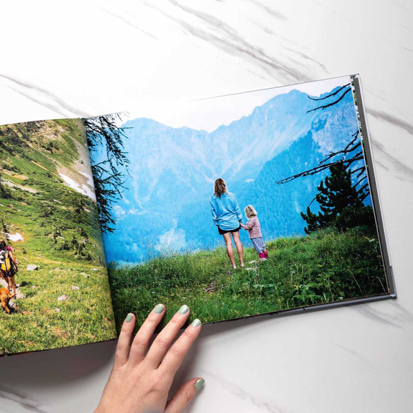 Open hardcover photo book displaying a full-bleed mountain landscape with a woman and child standing on grassy overlook, hand visible turning page.