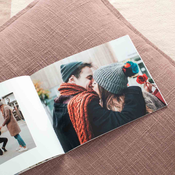 Open softcover photo book showing a couple in winter clothing sharing a kiss while holding a red rose, resting on a textured mauve pillow
