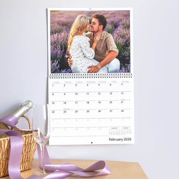 Wall calendar open to February 2026, showing a couple embracing in a blooming lavender field, with champagne and ribbon styled on a tabletop below