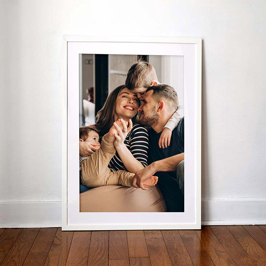 Framed and matted photo print of a smiling family cuddling on a couch, displayed against a white wall on a hardwood floor