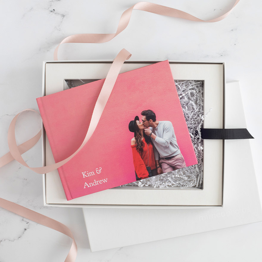 Layflat photo book with a pink cover featuring a couple kissing, placed in a white gift box with shredded paper and ribbon