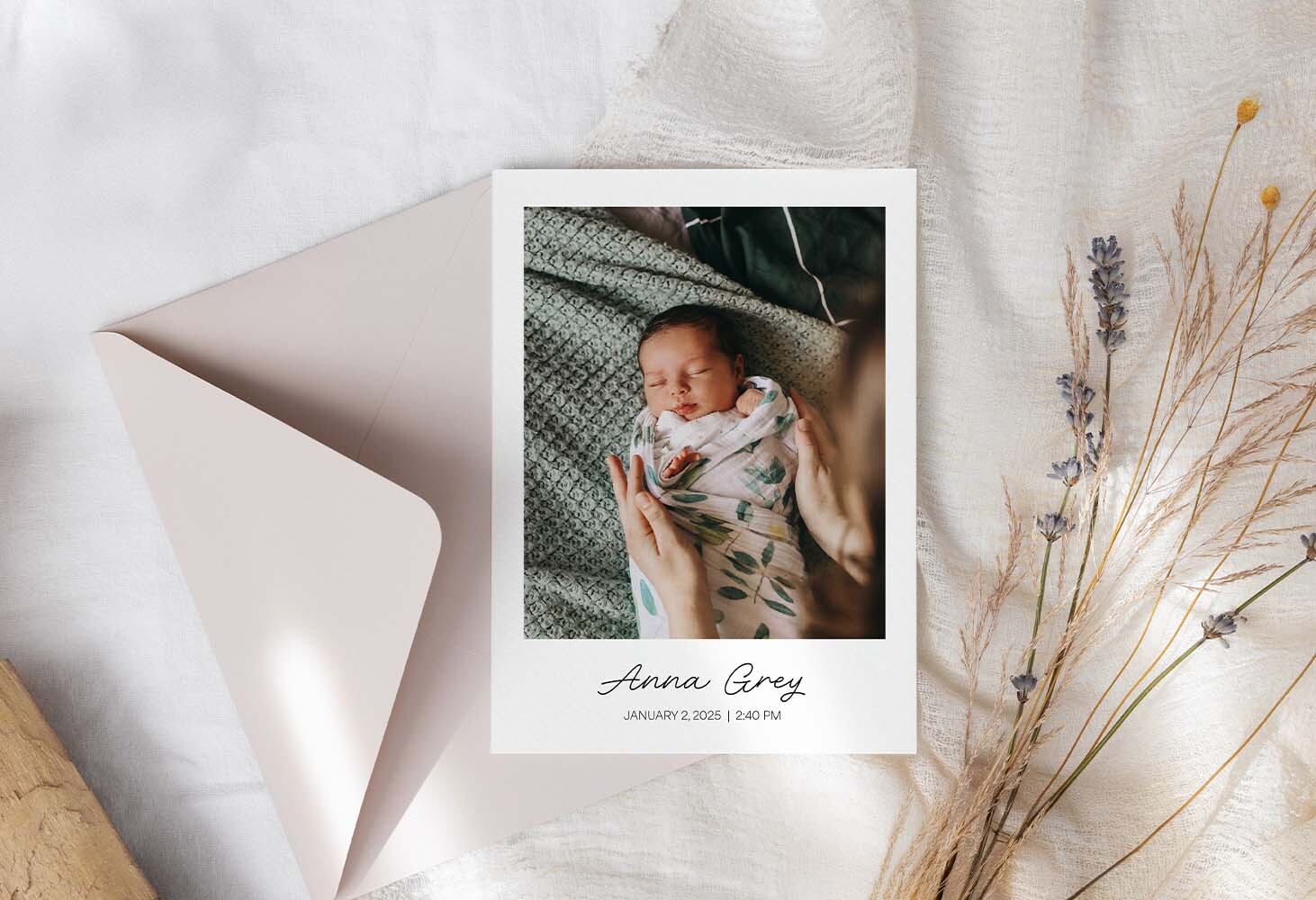 Flat newborn announcement photo card featuring a swaddled baby with printed name and birth details, styled with envelope and dried florals on neutral fabric background.