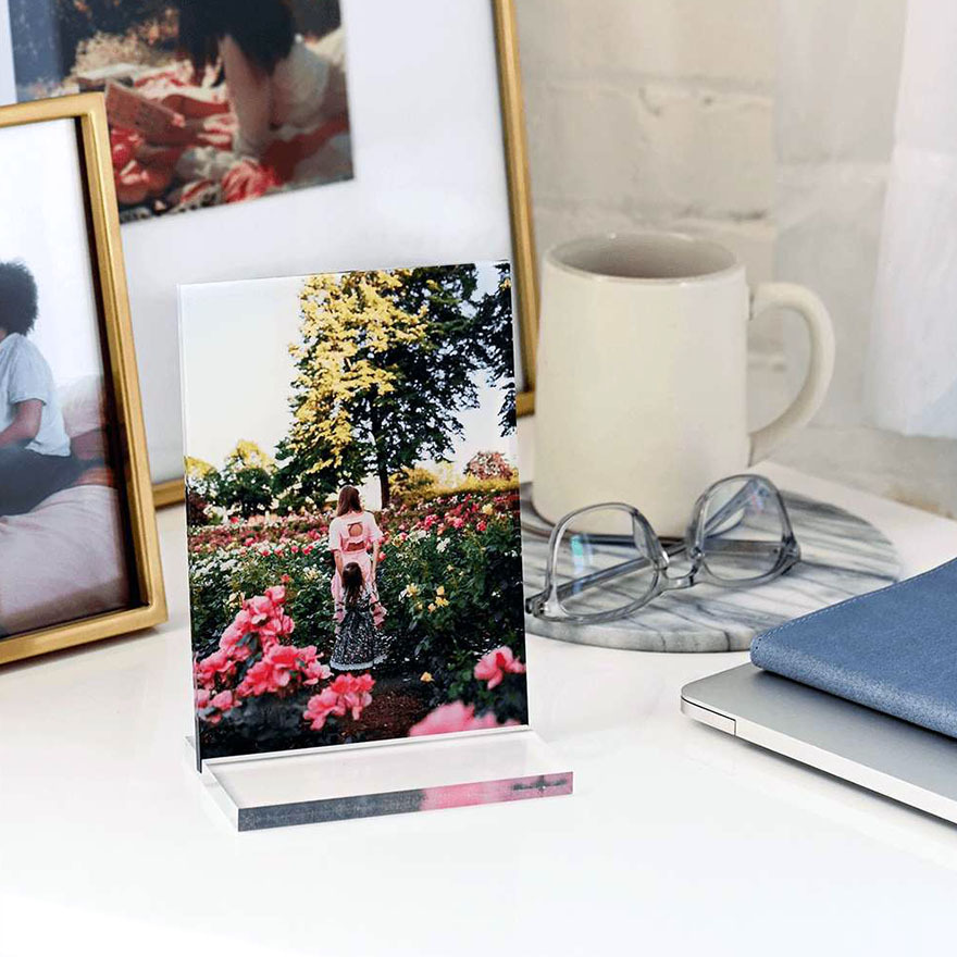 Clear acrylic desk photo print displaying a person standing in a flower garden, positioned on desk beside glasses, mug, laptop, and framed photos.