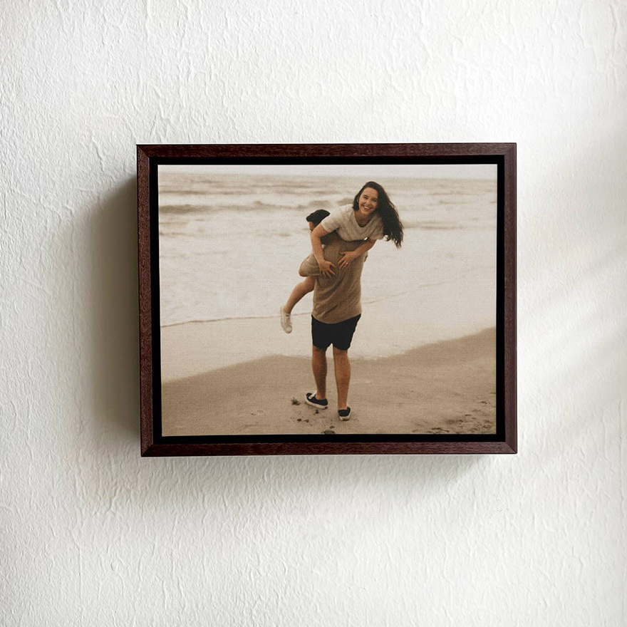 Framed canvas wall art showing a couple at the beach with one person lifting the other, displayed in a dark wood frame on textured wall.